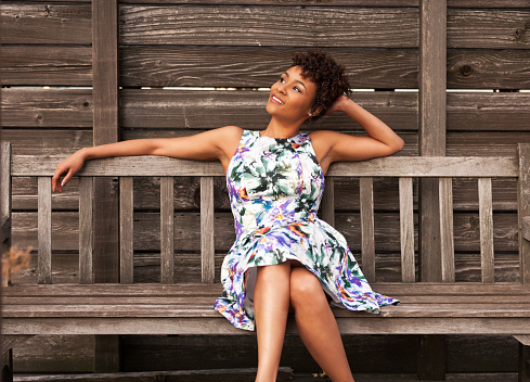 Attractive African American woman sitting on wooden bench wearing a sundress, enjoying the warm weather