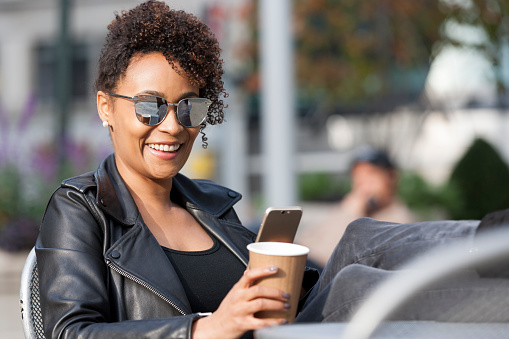 young African American woman enjoying a nice day outside with a cup of coffee on mobile phone at an outdoor cafe.
