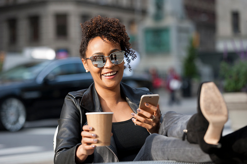 young African American woman enjoying a nice day outside with a cup of coffee on mobile phone at an outdoor cafe.