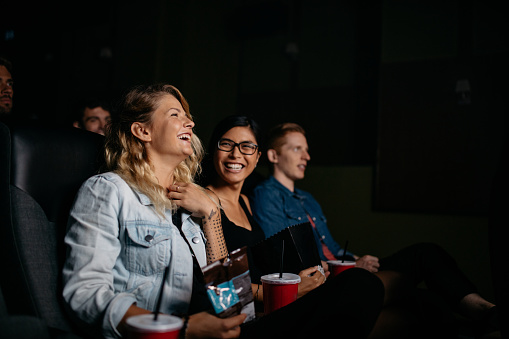 Group of young people watching movie in cinema. Friends sitting in movie theater with popcorn and drinks.