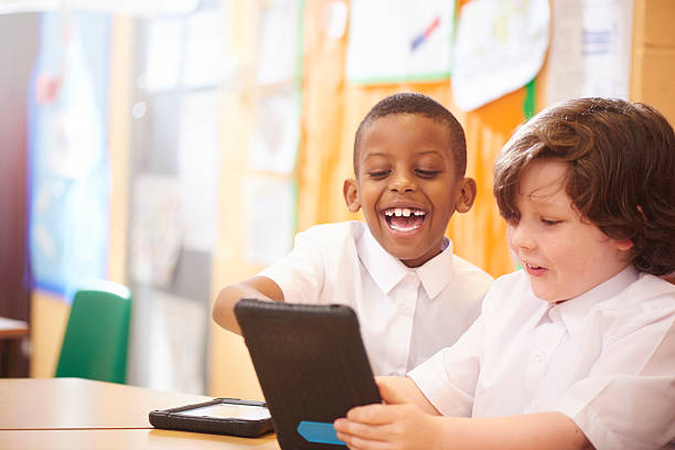 fun with technology two primary aged schoolchildren sit at their desks and look engrossed in the work that they are doing on their digital tablets. The little boy has an amazed expression whilst looking at his tablet and having fun . They are all wearing white school shirts school boy in uniform stock pictures, royalty-free photos & images
