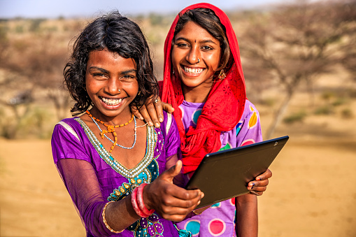 Happy Indian young girls sitting on a sand dune and using digital tablet in desert village, Thar Desert, Rajasthan, India.