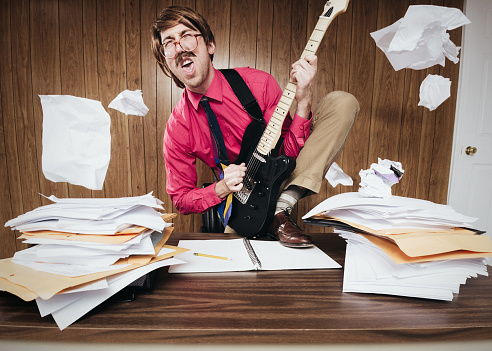 A white collar business man working in a retro 1980's style office stands on his desk piled with paperwork and documents, shredding some rock and roll on his electric guitar.  Papers are kick off of the desk from his antics.  GUITAR IS FULLY RELEASED.