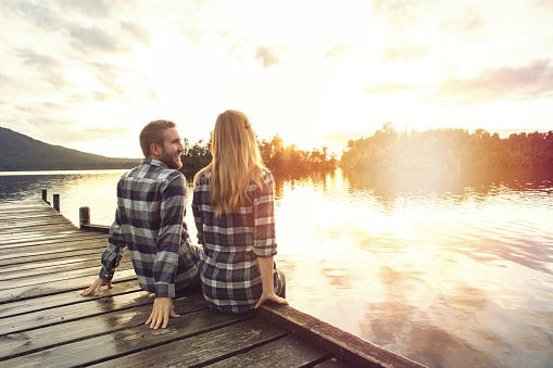 Cheerful young couple sitting on a lake pier chating. Lake Kaniere, New Zealand.