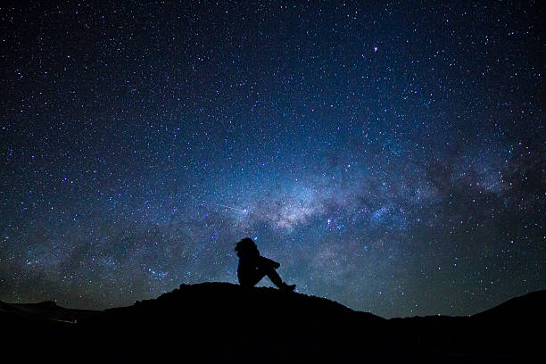Man's silhouette sitted, staring at the milky way Uyuni, Bolivia - October 30, 2016: Man's silhouette, sitted on a little hill, pointing to the sky, with the bright milky way as background. stare stock pictures, royalty-free photos & images