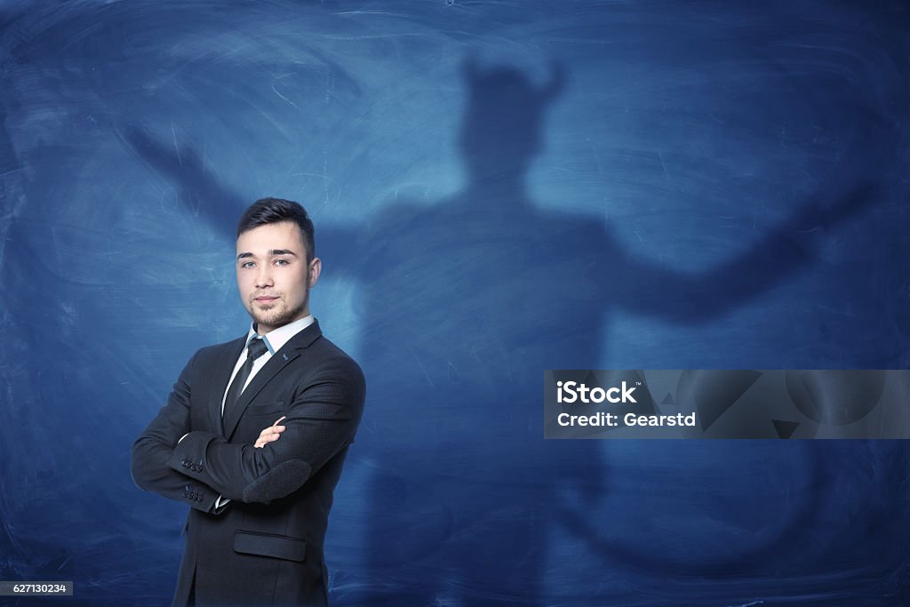 Businessman standing with hands across and his shadow on blue A businessman standing with his hands across and his shadow on a blue empty blackboard behind him with hands overspread, horns and a tail like a devil has. Self-confident behavior. Hidden feelings. Inner anger. Evil Stock Photo