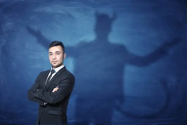 A businessman standing with his hands across and his shadow on a blue empty blackboard behind him with hands overspread, horns and a tail like a devil has. Self-confident behavior. Hidden feelings. Inner anger.