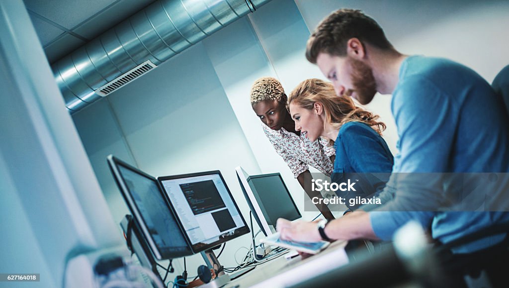 Application developers at work. Closeup side view of group of software developers working late at IT office. There are 3 people sitting side by side, a man and two women, African american and caucasian. Tilt shot. Technology Stock Photo