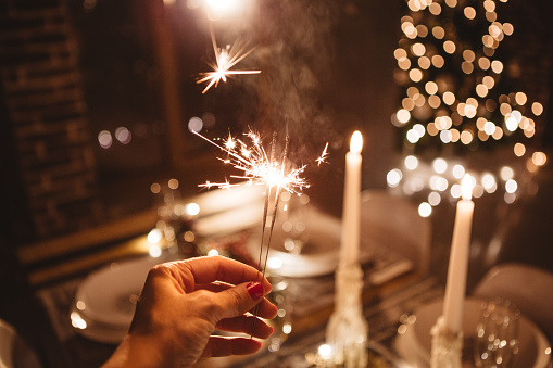 Shot of woman's hand holding sparklers in front of Christmas decorated table and tree. Evening or night.