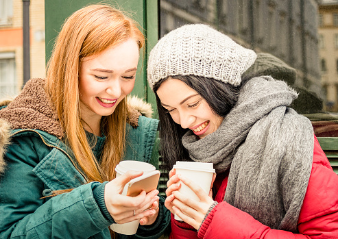 Happy girlfriends best friend having fun with coffee takeaway cup in autumn season - Friendship concept with joyful girls sharing time together with smartphone and winter clothes - Bright vivid filter
