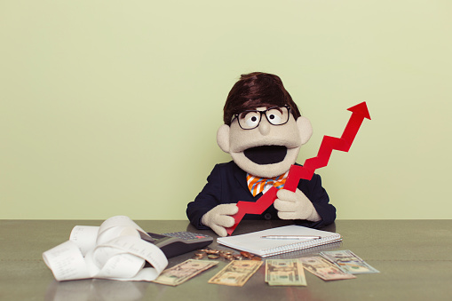 A puppet accountant counts his American dollars as his business is booming. He is smiling while holding a red line graph and ready to grow your business. Dressed in business suit and bow tie. Retro styled.
