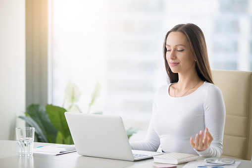 Young woman near the laptop, practicing meditation at the office desk, in front of laptop, online yoga classes, taking a break time for a minute, healing from paperwork and laptop radiation