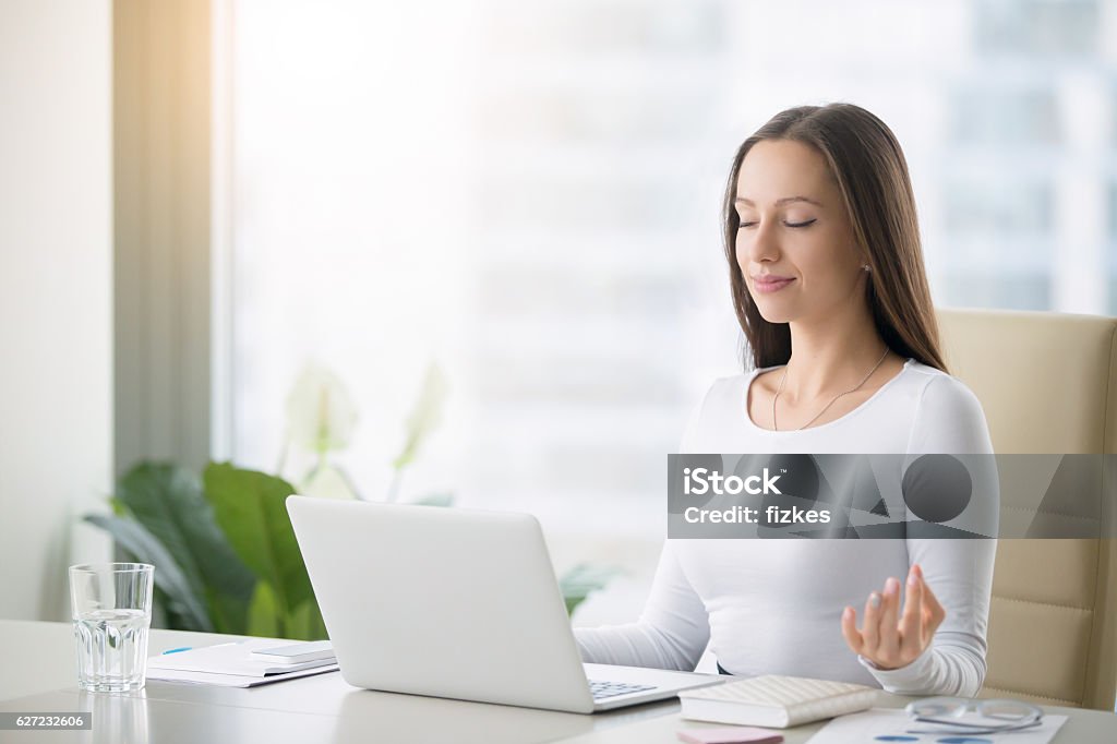 Young woman practicing meditation at the office desk Young woman near the laptop, practicing meditation at the office desk, in front of laptop, online yoga classes, taking a break time for a minute, healing from paperwork and laptop radiation Meditating Stock Photo
