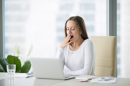 Young business woman yawning at a modern office desk in front of laptop, covering her mouth out of courtesy, chain reaction, drowsiness, unable to deal with boring job, monday after cool weekends