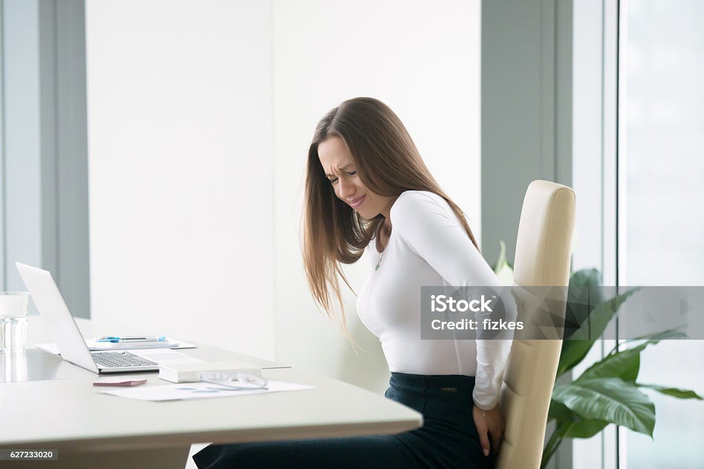 Young businesswoman with a backpain Profile portrait of an irritated young businesswoman woman at the office, feeling her back tired after working at laptop, uncomfortable chair, feeling severe back ache, itching, difficulty sitting Hemorrhoid Stock Photo