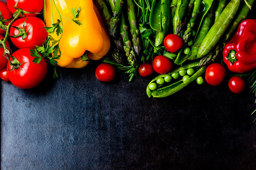 Food background - raw organic vegetables, fresh ingredients for healthily cooking on black background - bell pepper, green pea, asparagus, tomatoes, rosemary. top view. Vegetarian or healthy food