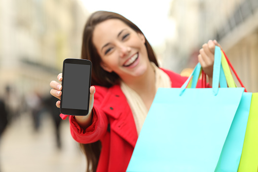 Front view of a shopper holding blank shopping bags showing to the camera a smart phone screen with an urban background