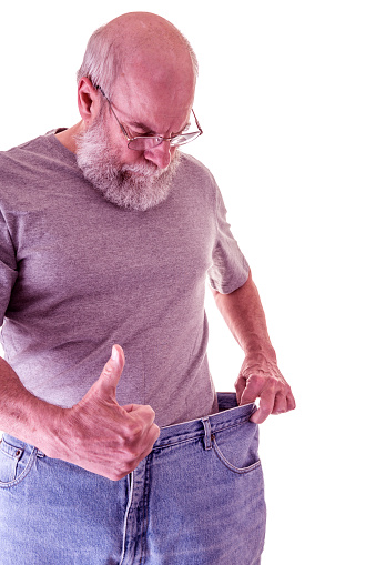A proud, trim, senior adult man is giving the thumbs up hand signal. He is showing off - stretching and pulling out the waistband of his old jeans - now way too big for him to wear - to show how well his healthy living fitness program is going, and to display how much weight he has lost.