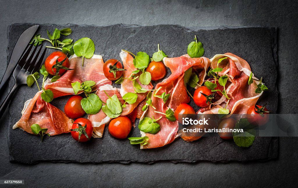 Salad with ham jamon serrano, cherry tomatoes, arugula, slate board Salad with ham jamon serrano, cherry tomatoes, arugula on black stone slate board, grey background. Top view Parma Ham Stock Photo