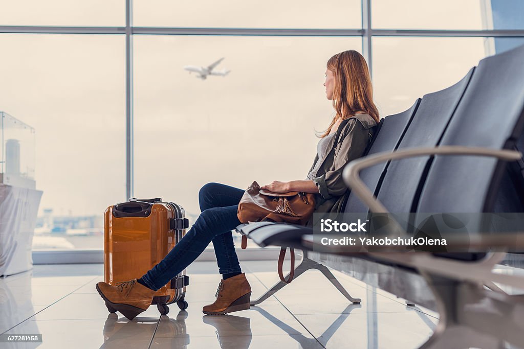 Smiling girl waiting for boarding Start of her journey. Beautiful young woman looking out window at flying airplane while waiting boarding on aircraft in airport lounge Airport Stock Photo