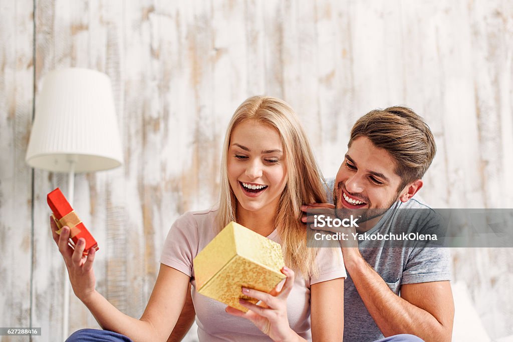 Cute loving couple celebrating anniversary What a wonderful gift. Surprised young woman is opening box and smiling. Man is sitting and embracing her with love Gift Stock Photo