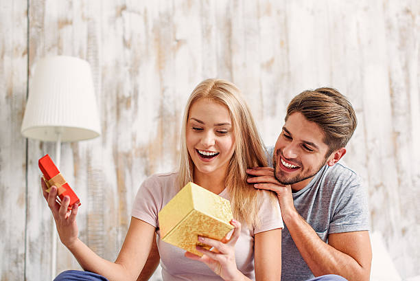 Cute loving couple celebrating anniversary What a wonderful gift. Surprised young woman is opening box and smiling. Man is sitting and embracing her with love gift for girlfriend stock pictures, royalty-free photos & images