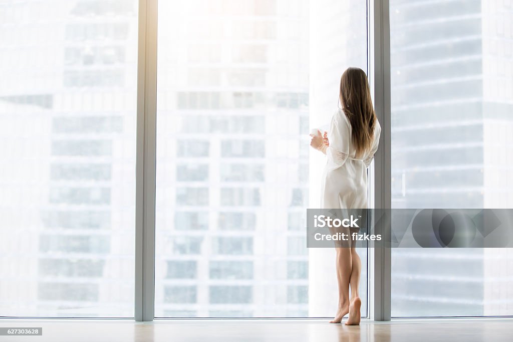 Young woman near the floor window Young woman, wearing dress gown looking through floor window, waiting for a busy husband, dreaming of a job in big city, first day in unknown town, ready to conquer the capital. Rear view, full length Women Stock Photo