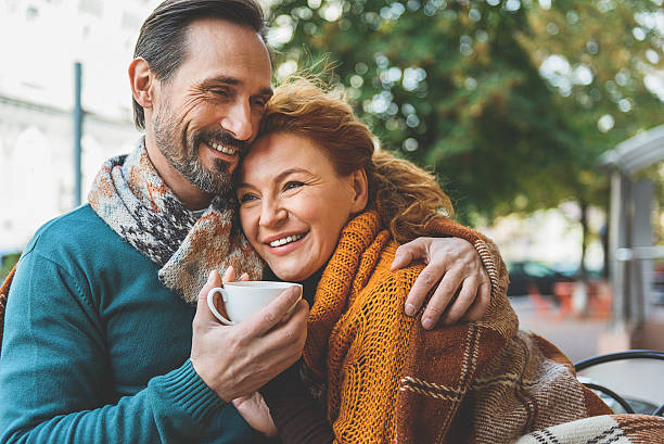 Pretty senior lovers hugging with joy Happy loving couple is dating in cafeteria outdoors. They are sitting covered by blanket and laughing. Man is embracing woman and holding cup mature couple stock pictures, royalty-free photos & images
