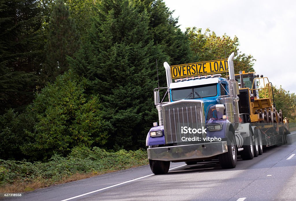 Oversize load rig semi truck with step down flat bed The big powerful rig semi truck with a sign Oversize load on the cab roof and special flat bed trailer which transports a large construction equipment on a straight interstate road Trucking Stock Photo