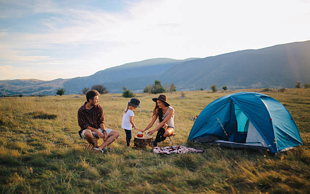 Young people camping with a baby girl Landscape in Dinaric Alps, mountain range in Southern Europe, on the border of Croatia and Bosnia. Young couple is camping with a baby girl child. They are sitting in the in front of a small tent, enjoying a beautiful view over the valley under the mountains, watching the horizon as the sun sets, playing games and having fun in the nature outdoors. They're dressed in fashionable hippie clothes, enjoying freedom in the nature, wearing hats and having a coffee in a small stove top espresso pot. family camping stock pictures, royalty-free photos & images