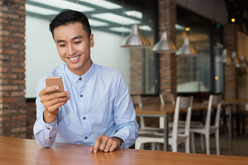 Closeup portrait of smiling young Asian man using smartphone and sitting at empty table with blurred cafe interior in background