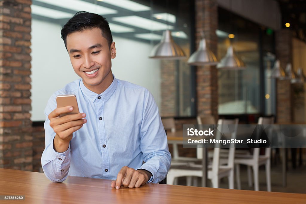 Smiling Asian Man Using Smartphone at Cafe Table Closeup portrait of smiling young Asian man using smartphone and sitting at empty table with blurred cafe interior in background Asian and Indian Ethnicities Stock Photo