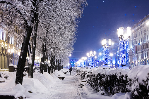 winter city street with trees and benches covered in snow and lamp posts decorated with christmas lights