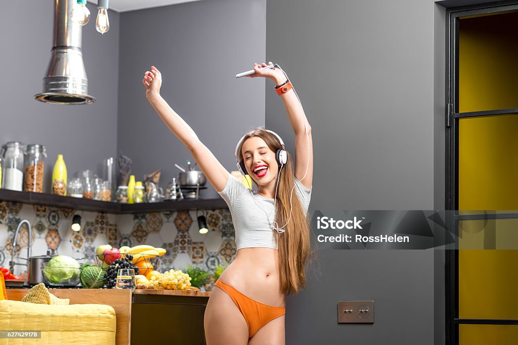 Woman in the kitchen Lifestyle portrait of a young sports woman listening to the music with phone and headphones in the modern kitchen interior full with fruits and vegetables Kitchen Stock Photo