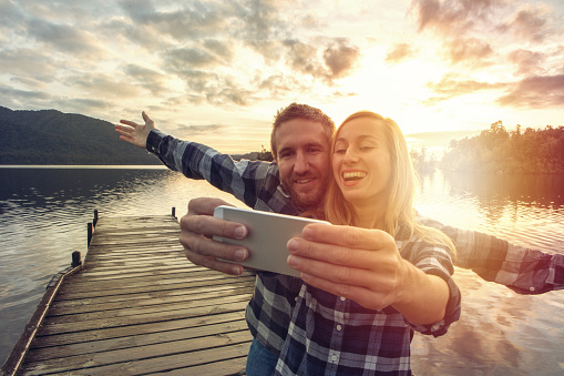 Young couple on pier above lake take a selfie portrait using smart phone, spectacular landscape on the background.