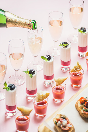 Various snacks, brushettas, gazpacho shots, desserts with berries and champagne pouring to glasses on corporate event, christmas, wedding celebration over pastel pink background, selective focus