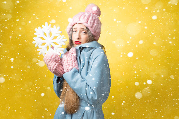 Confused and unhappy woman in colorful winter clothes holding a snowflake on the yellow background