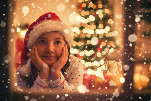 Christmas Eve,beautiful little girl with Santa hat sitting in living room in warm holiday atmosphere and she looking through window,while waiting for Santa 