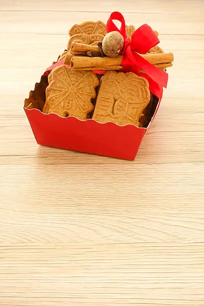 Red tray full of spiced biscuits with almonds, Spekulatius mit Mandeln, decorated with a red Christmas bow, cinnamon sticks, cloves and muscat. Backlit, light brown wooden background and lots of copy space.