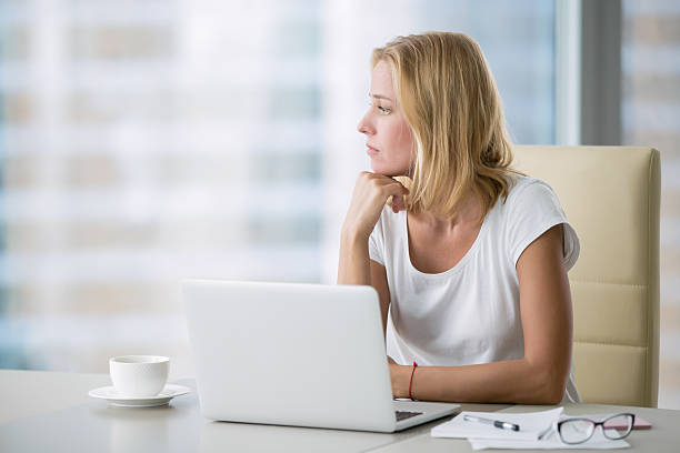Yong woman with laptop looking at the window Young attractive woman at a modern office desk, working with laptop, looking at the window, thinking about a post, full-time blogger, seeking for inspiration, help to be productive, updating computer drudgery photos stock pictures, royalty-free photos & images