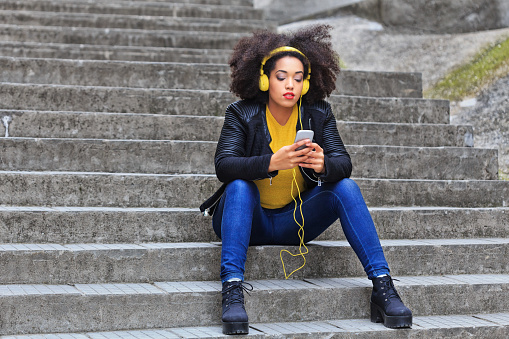 Pensive young woman sitting on stairs and listening music on smart phone. Yellow headphones on head, wears black jacket, yellow blouse and blue jeans. Stone stairs on background.