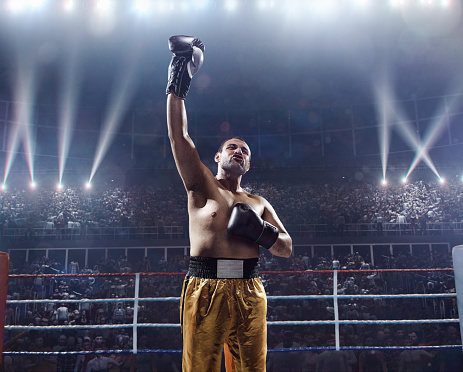 A picture of a winner boxer with his arms to the air. Sportsman is on boxing ring with bleachers full of people