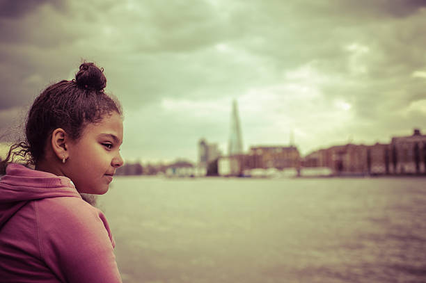 Stock photo of a little girl aged 10 years old who is sitting on the bank of the river Thames. She is looking pensive with her gaze lost to the horizon. One can see the Shard and Tower Bridge in the background.