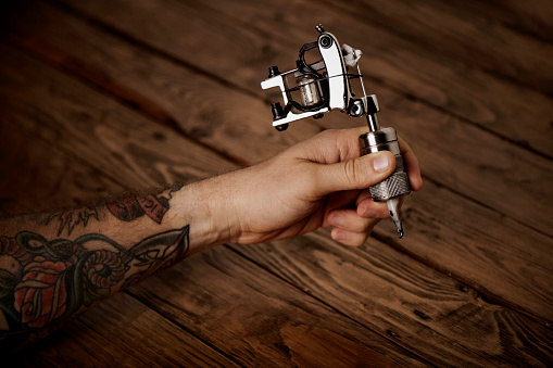 Brutal tattooed man's hand with a custom made induction tattoo machine above rustic wooden table