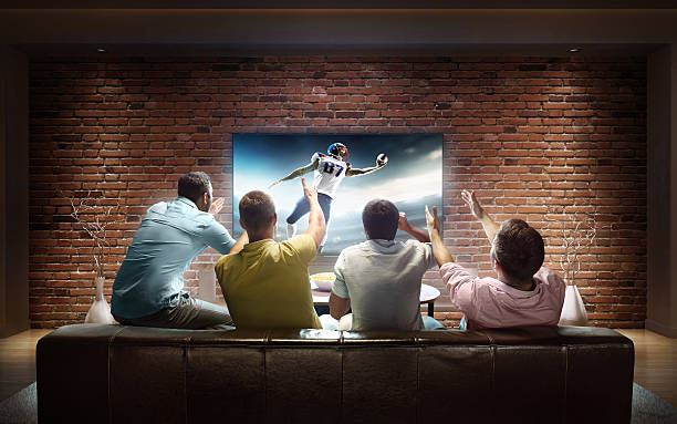 Students watching American football game at home :biggrin:A group of young male friends are cheering while watching American football game at home. They are sitting on a sofa in the modern living room faced to a big TV set on the front wall. watch stock pictures, royalty-free photos & images