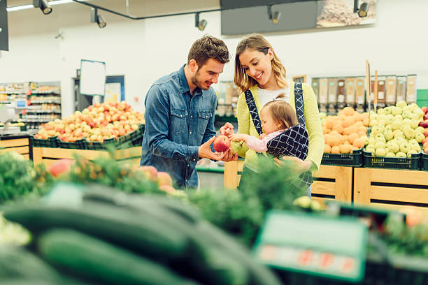 Young Family With Their Baby Daughter Groceries Shopping Young family groceries shopping. Mother carry their baby daughter in baby carrier and father choosing apples. 3 6 months stock pictures, royalty-free photos & images