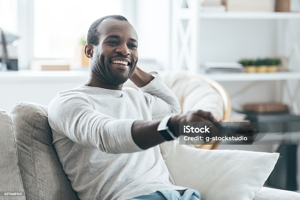 Watching TV at home. Handsome young African man watching TV and smiling while sitting on the sofa at home Watching TV Stock Photo