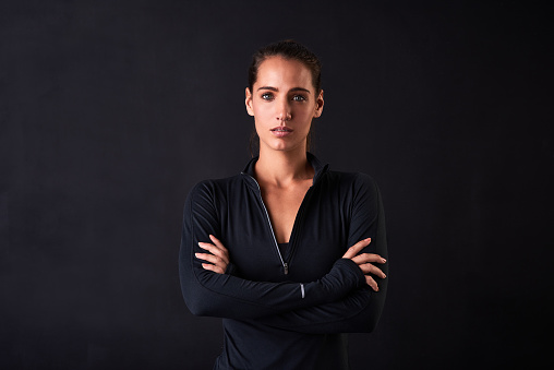Studio portrait of a young woman in gym clothes standing with her arms crossed against a dark background