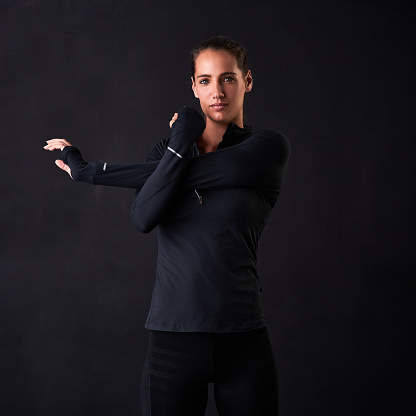 Studio portrait of a young woman in gym clothes stretching her arms against a dark background