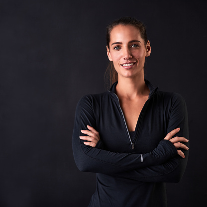 Studio portrait of a young woman in gym clothes standing with her arms crossed against a dark background